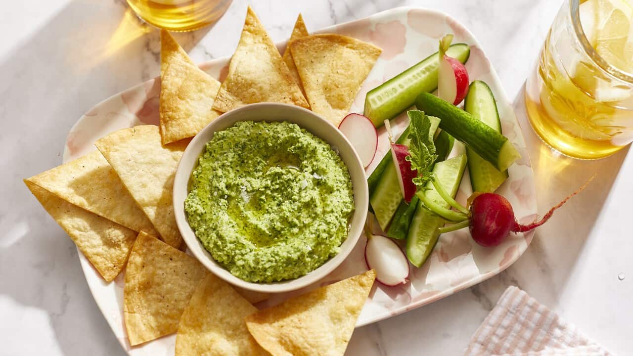 A bowl of vibrant green dip sits on an oblong tray, with golden tortilla chips and fresh vegetable crudite. Two drinking glasses with a golden liquid in them sit alongside.