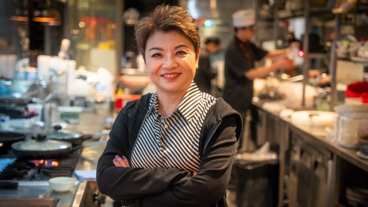 A woman in a striped shirt and black jacket standing with her arms folded inside a restaurant kitchen.