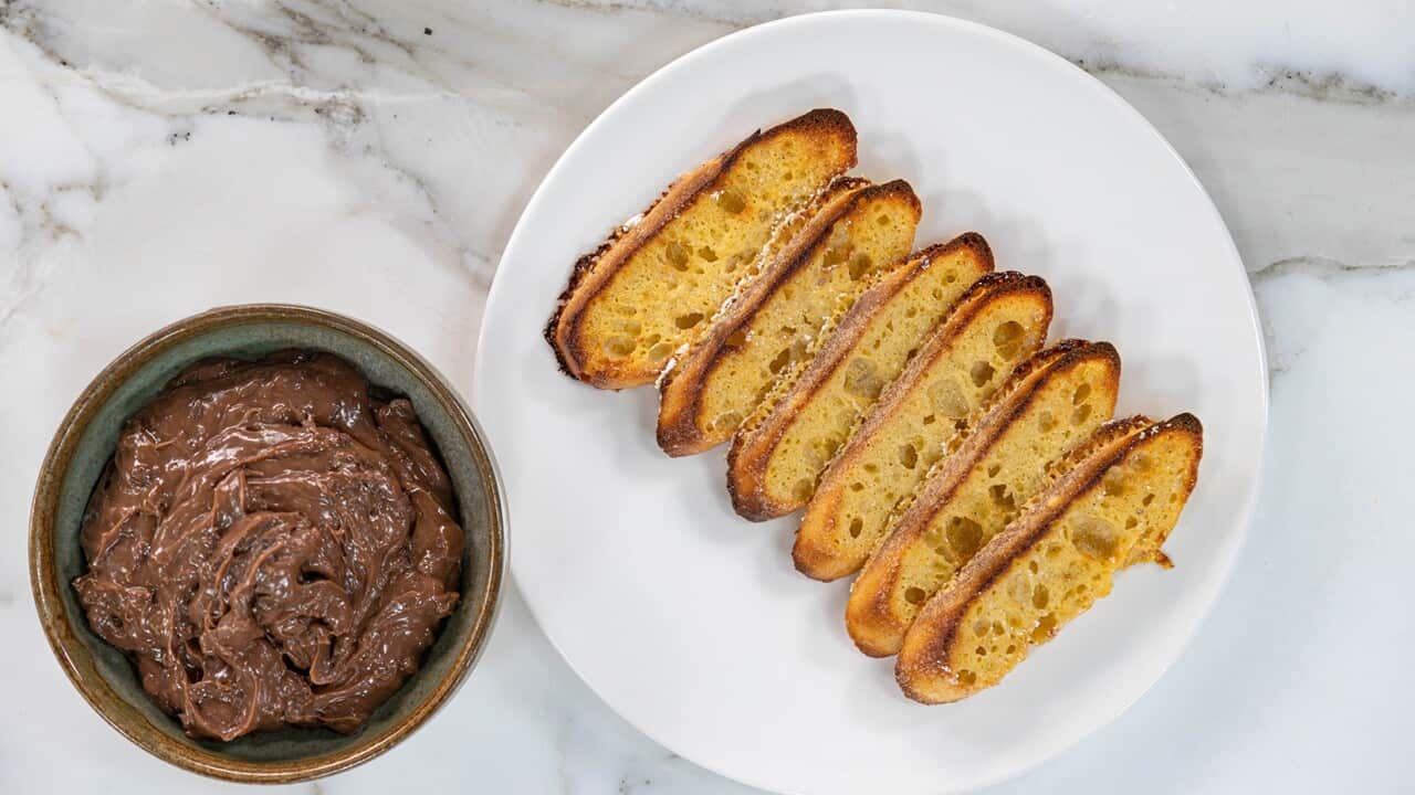 Six long toasted baguette slices sit on a round white plate. The edges of the bread appear to have a little sugar here and there. To one side sits a small dish with a rich brown spread in it.