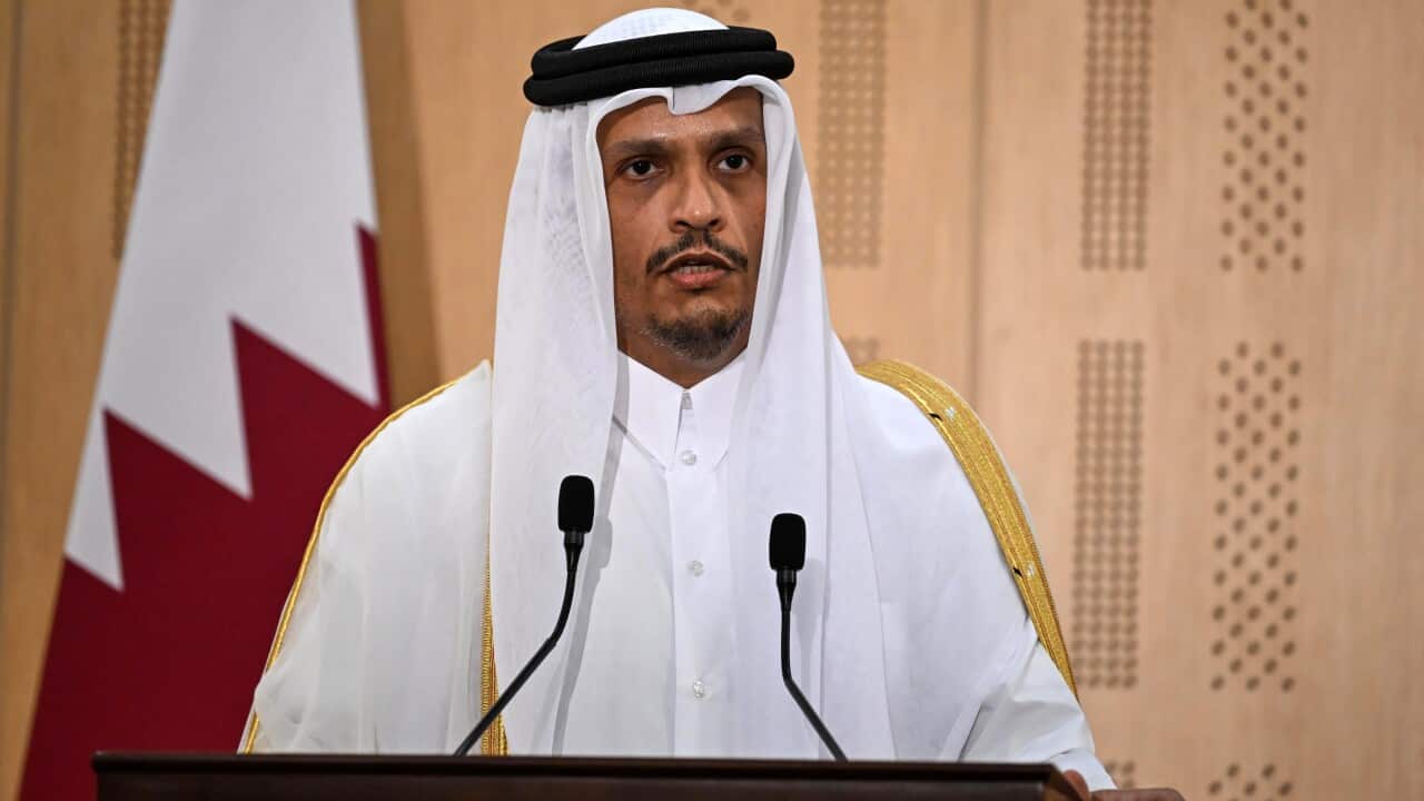A man in a thobe is speaking at a podium in front of a Qatari flag.