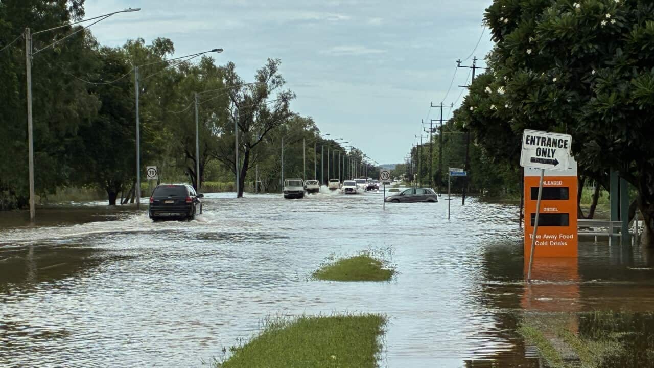 A photo of a flooded main street in Katherine. Cars are still able to pass through the water, though it appears one small car has become stranded in a deeper patch. A nearby petrol price tower sits half submerged.