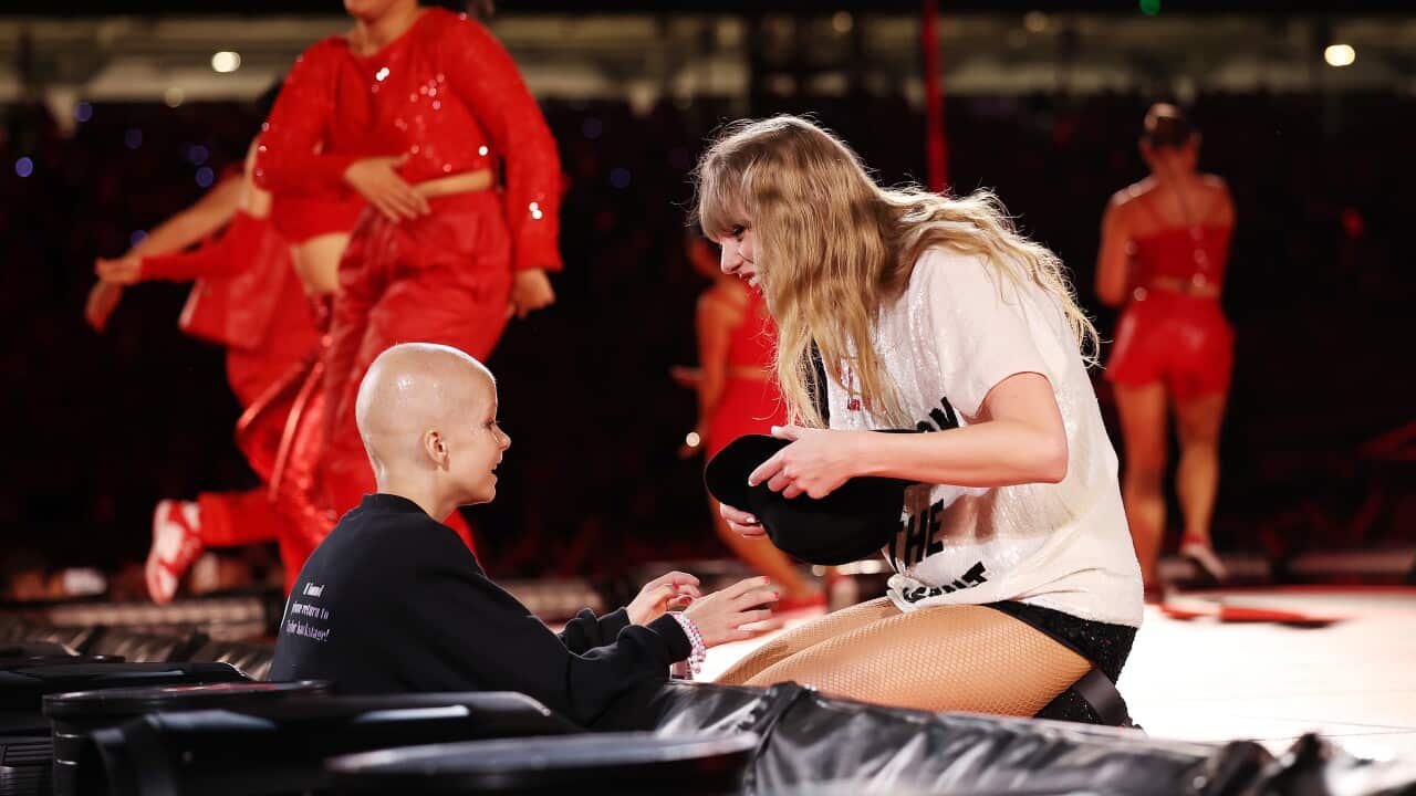A young girl dressed in black and with no hair stretches her hand towards a woman with long blonde hair on stage, who is holding a black hat and kneeling towards the girl.