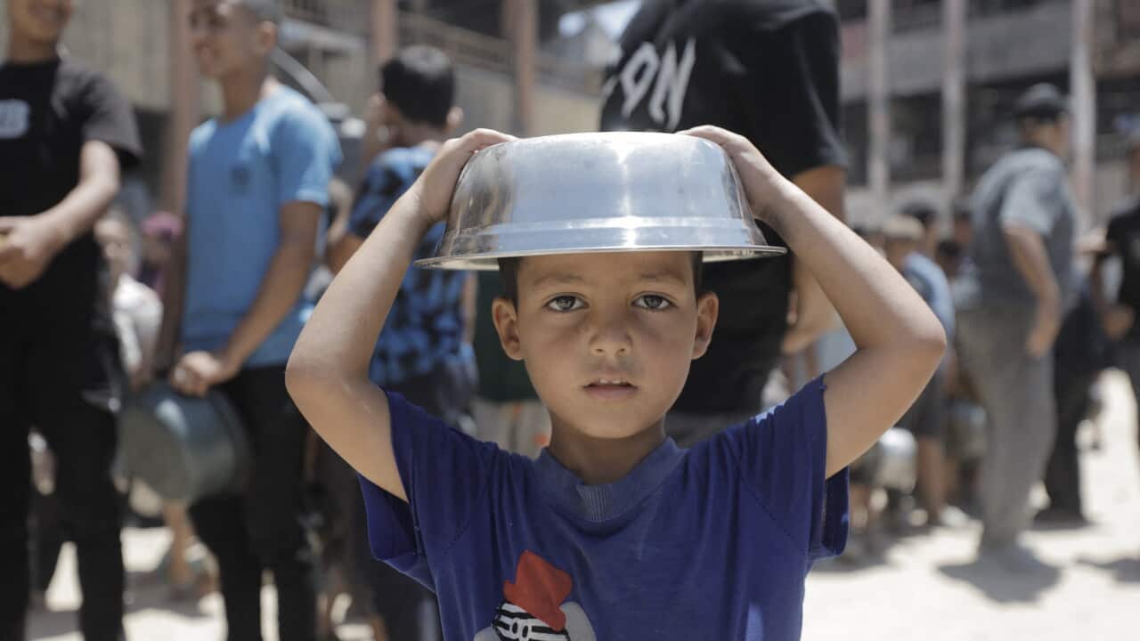 A boy in Gaza wearing a blue shirt holds an empty cooking pot over his head, behind him a line of people wait for food
