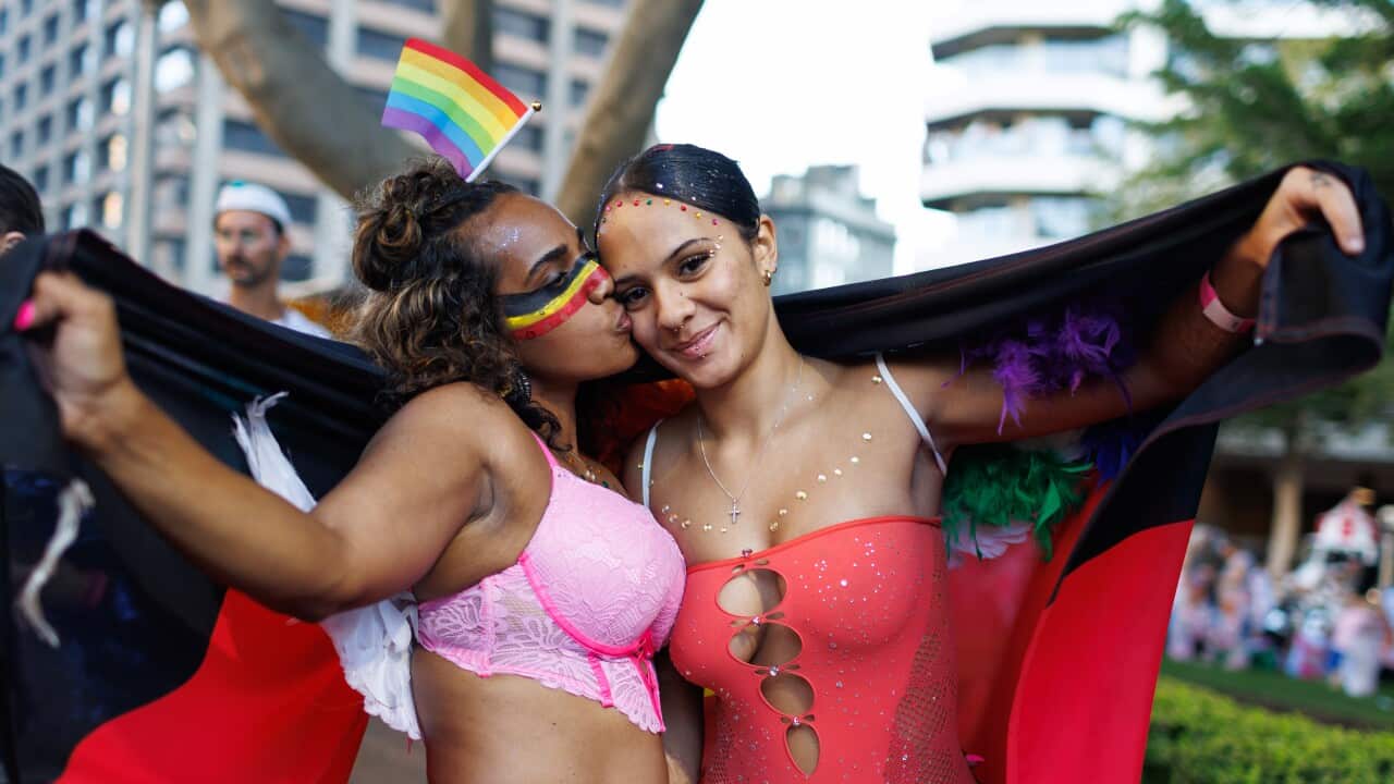 A person in pink lingerie kisses another on the cheek. They are holding a black, yellow and red Aboriginal flag over their shoulders.