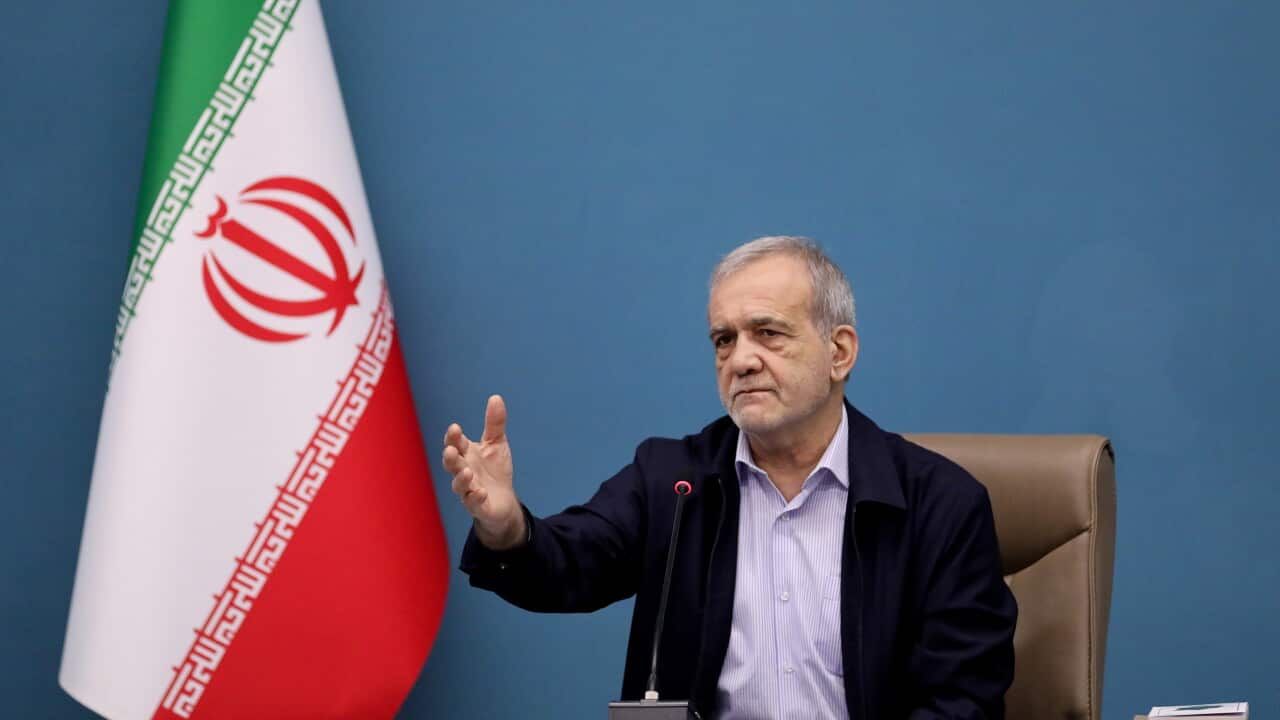 An older Persian man sitting at a desk with his right arm in front of him. An Iranian flag is beside him.