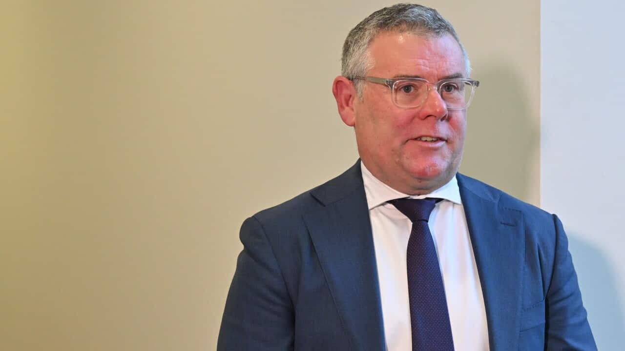 A person in a blue blazer and tie and white shirt standing in front of a light yellow background.