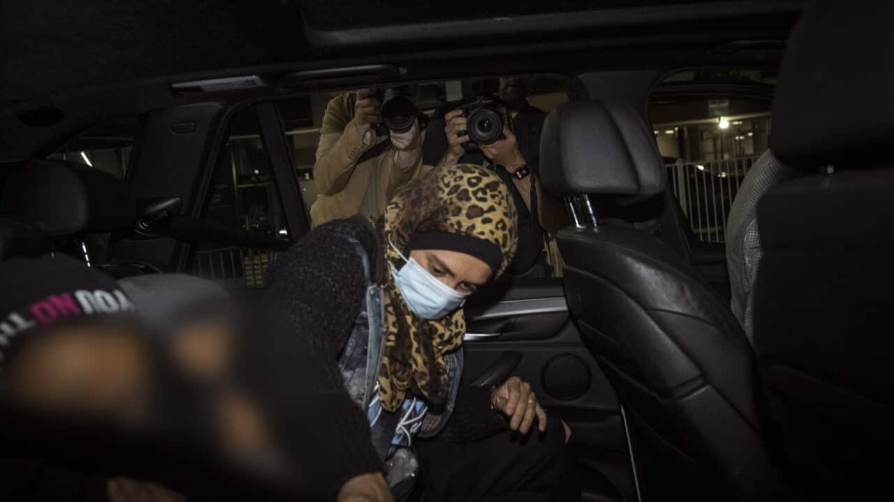 A woman wearing a headscarf and facemask gets into a car while members of the media take her photo.