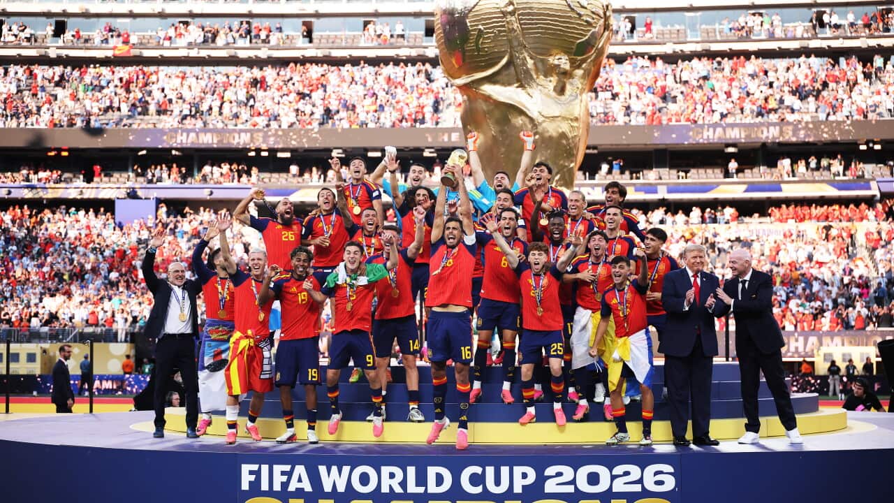 The Spanish national men's football team on a stage after winning the FIFA World Cup final. They are holding up a golden trophy in celebration. Behind them is a large FIFA trophy statue, and a crowd of people in a football stadium.