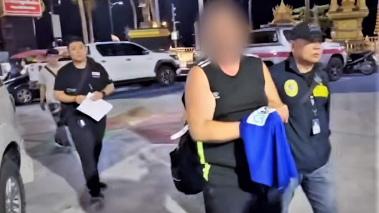 A man with his face blurred is walked through a street in Thailand by two police officers.