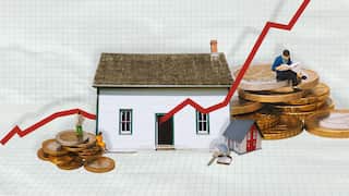 A graphic image shows a white house surrounded by stacks of coins, silhouettes of people and keys. A red graph line goes through the image, with grid paper in the background.