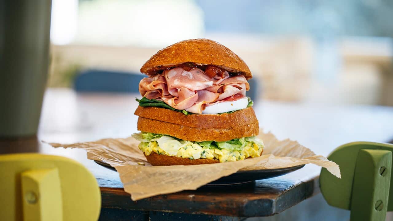 A double sandwich - a bun split into four 'slices' and filled with sliced meat in the top half and an egg-mayo mixture in the bottom - sits on a plate on the edge of a table.