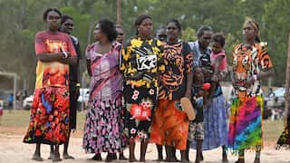 A group of nine indigenous women stand together in a sandy park. They are dressed in colourful clothing
