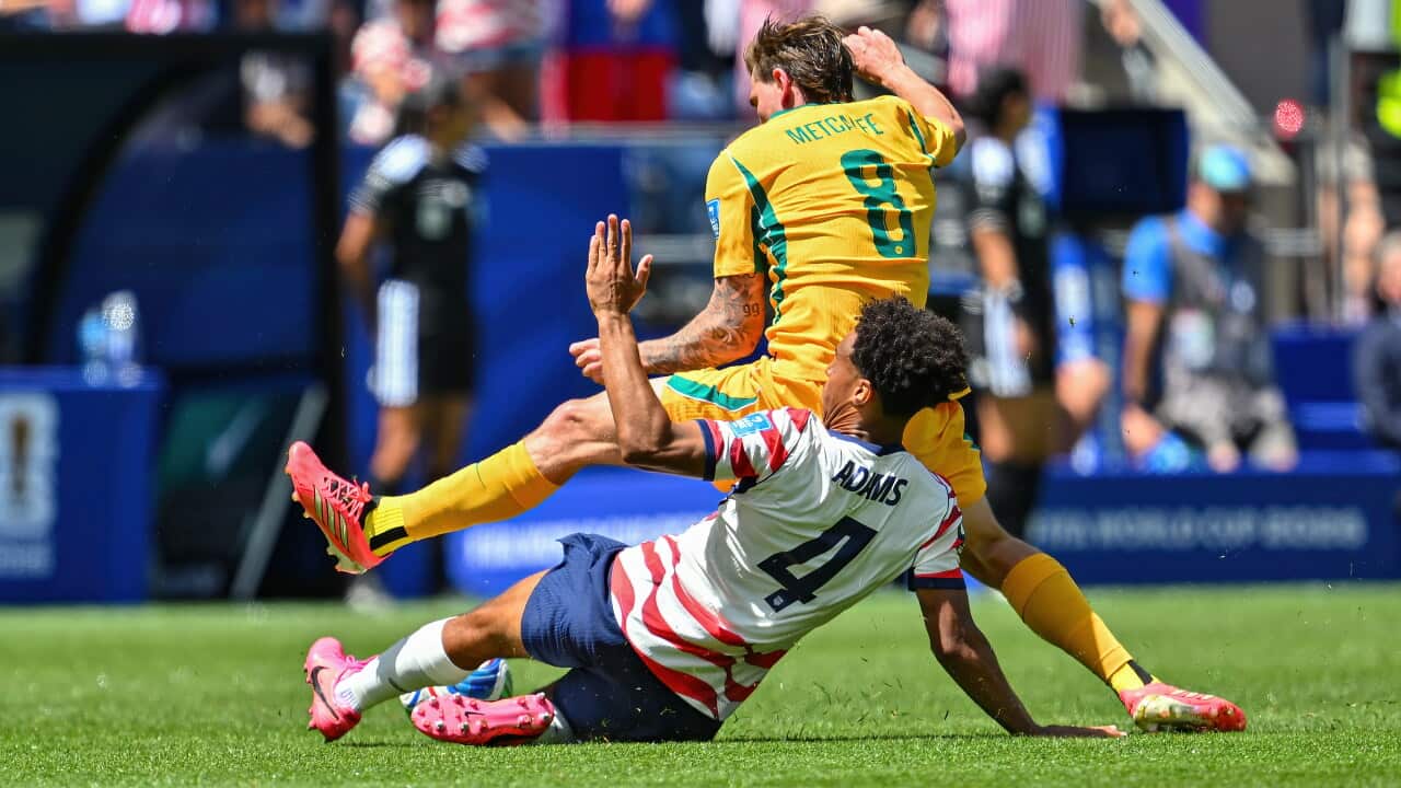 An action-filled, full shot of a soccer match showing a dynamic tackle between two players on a vibrant green field. A player from the US team wearing a white jersey with red stripes and the name "ADAMS" and number 4 on the back is sliding on the grass. He is tangled with an Australian player in a bright yellow kit with the name "METCALFE" and number 8 on the back, who is mid-air and losing his balance. Bits of grass and dirt are kicked up around them, and the background shows a blurred stadium crowd under bright, sunny daylight.