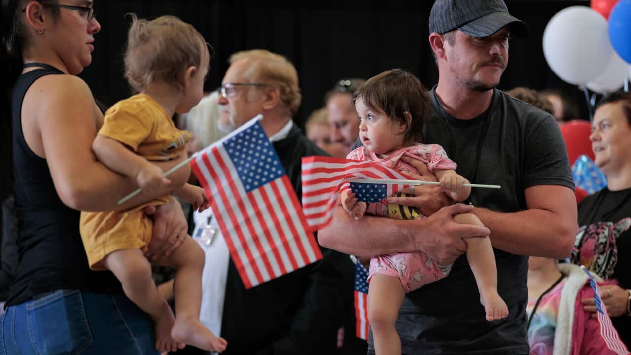A man and a woman, each holding a child who is holding an American flag in their hands, are standing. A group of people are standing behind them.