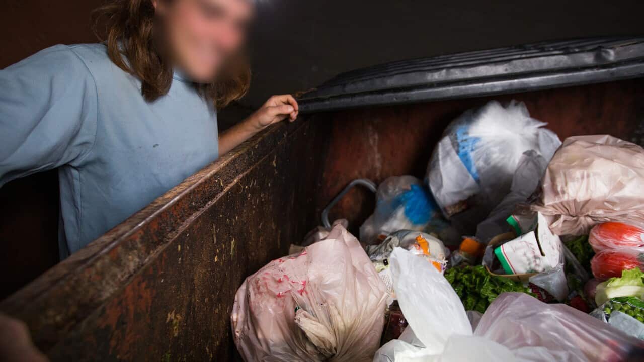 A man with his face blurred, looking into a skip bin filled with food