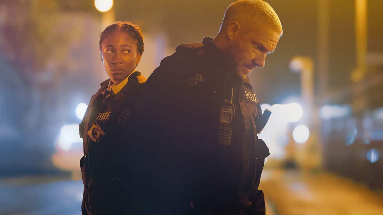 Two police officers, a man and a woman, stand in a dim city street. The man is slightly hunched over.
