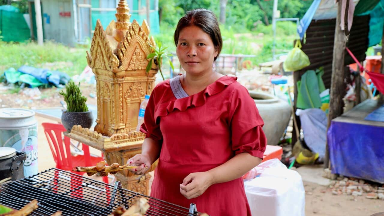 A Cambodia woman in a red dress stands in an open-air kitchen holding meat skewers over a grill with a golden spirit house in the background