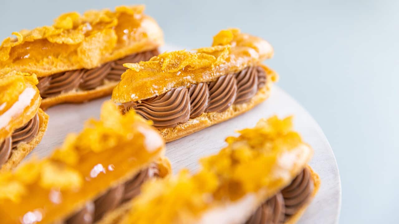 Five eclairs, filled with piped chocolate cream, sit on a round plate. Each eclair is coated with a toffee glaze and cornflake topping.