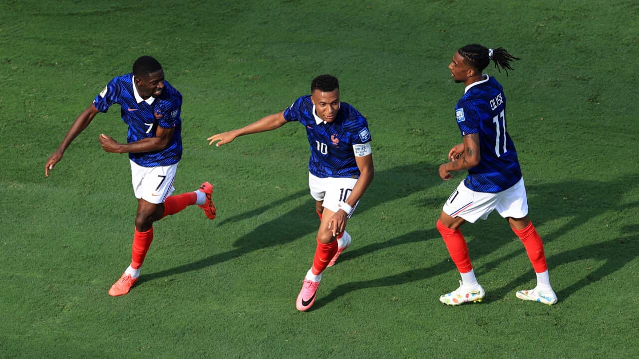 Three French footballers in blue jerseys, white shorts and red socks celebrate on the pitch after scoring, running toward each other with smiles and arms outstretched during a World Cup knockout-stage match.