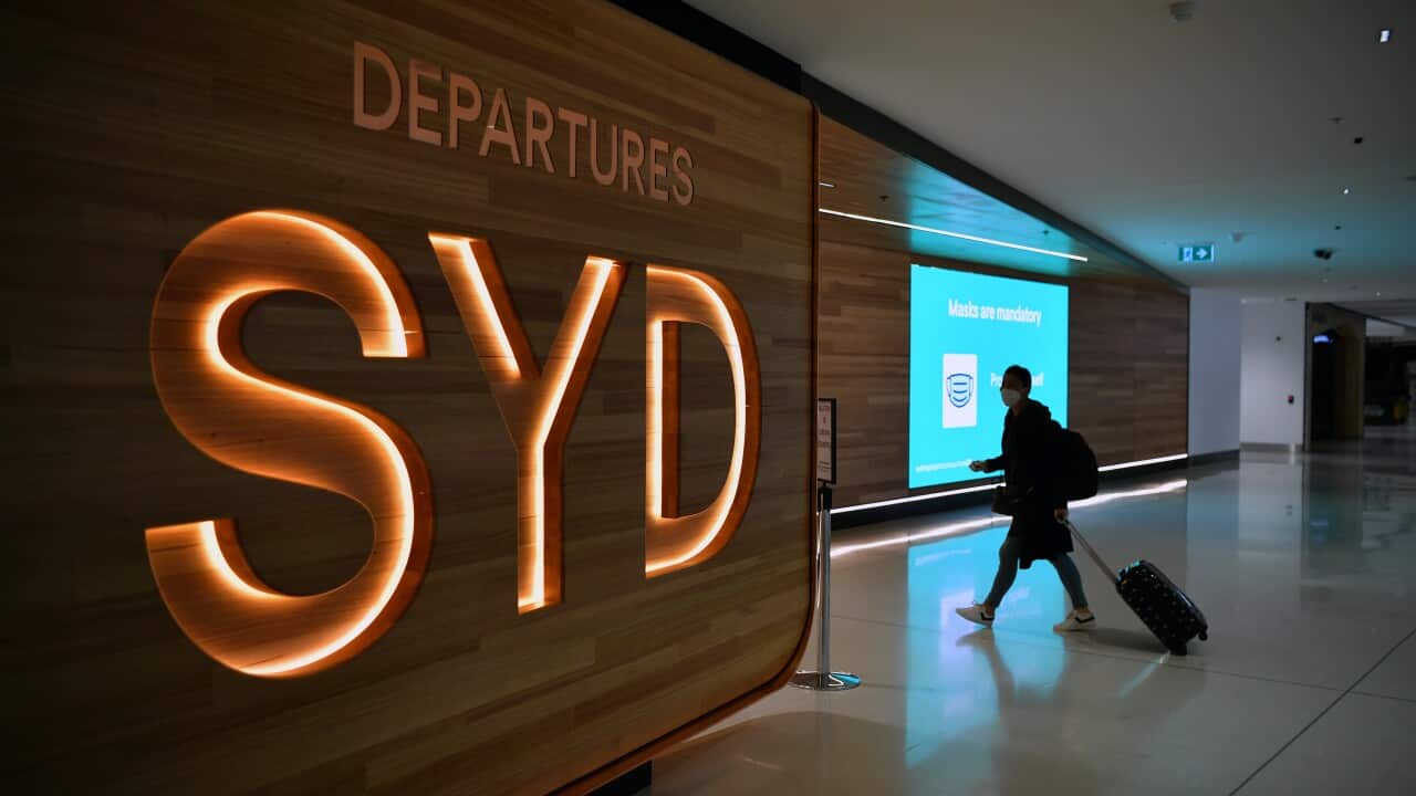 A person wheeling a suitcase in front of an LED sign reading SYD