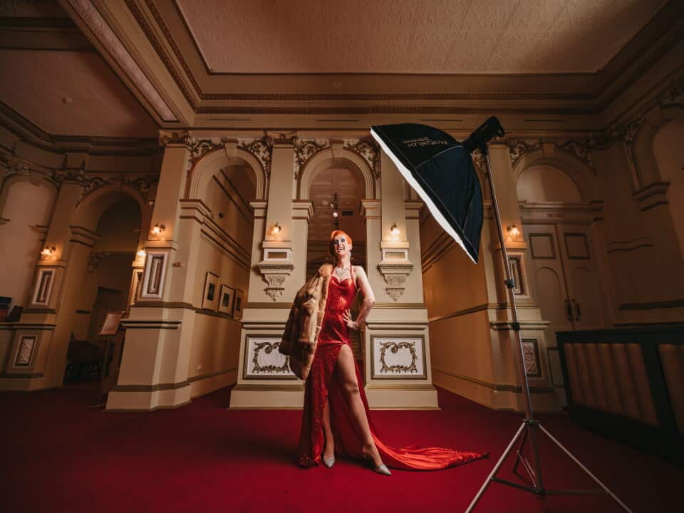 Cougar Morrison is a fabulous drag queen standing, laughing, on a red carpet in a sequin red gown with her grandmother's old fur coat draped over one shoulder