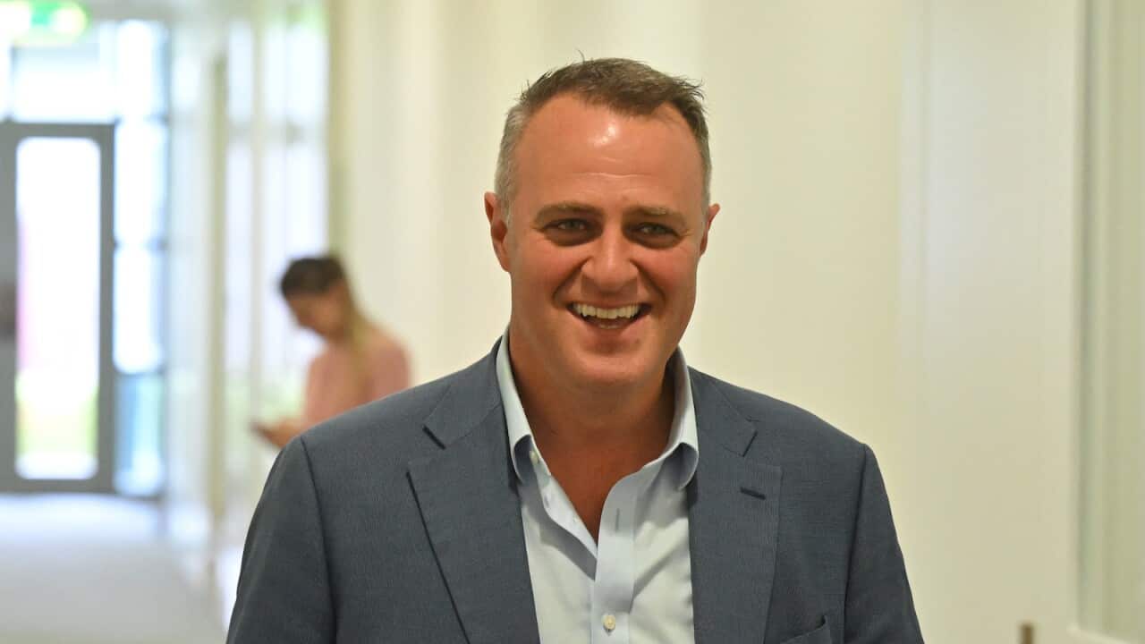 A man in a dark blue blazer and light blue shirt walking down a hallway, smiling.