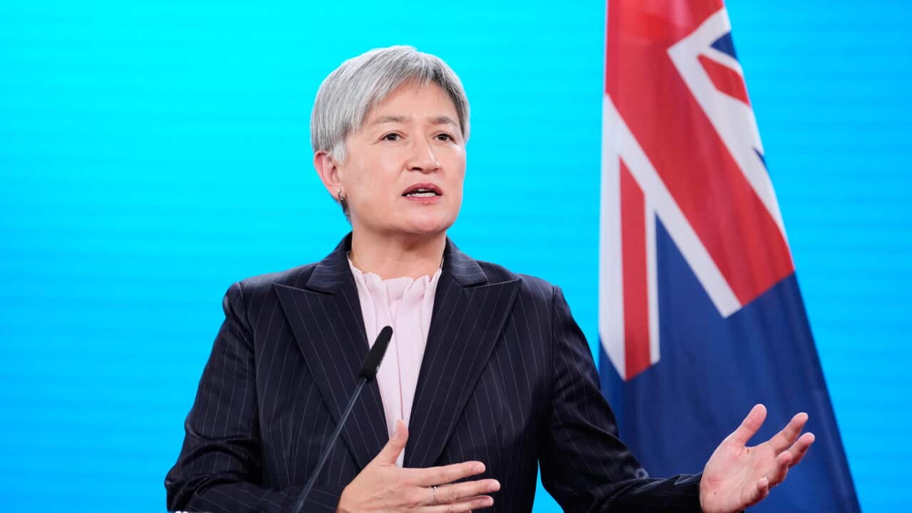 Penny Wong speaks into a microphone mounted on a podium, gesturing with her hands against a bright blue background featuring the Australian flag.
