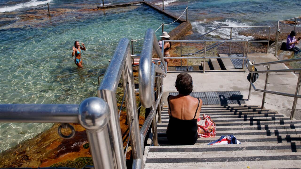 A woman in a black one-piece bathing suit sits on the steps leading to an ocean swimming pool.