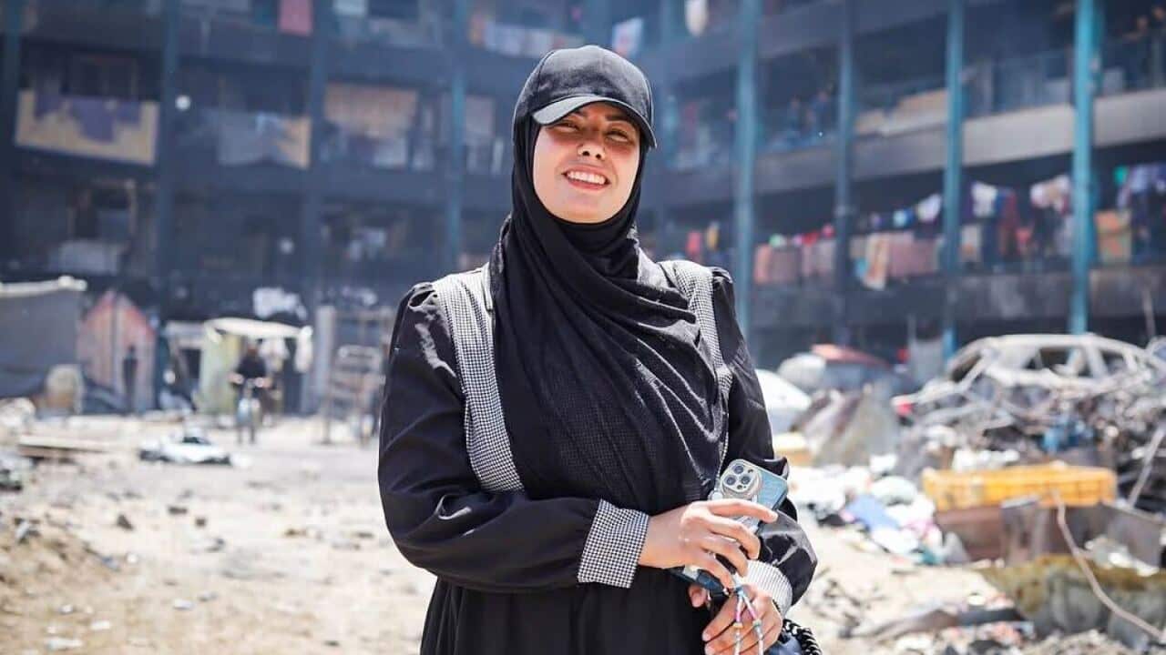 A woman wearing a black cap and hijab holding a phone standing in front of a damaged building and debris