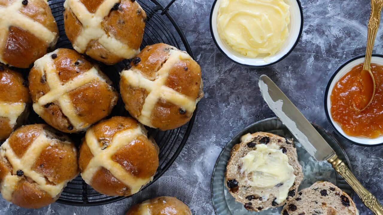 Close-up image of homemade Easter hot cross buns on cooling rack, sliced and buttered spiced bun on plate, dish of apricot jam, butter dish and knife, mottled grey background, home baking concept, elevated view