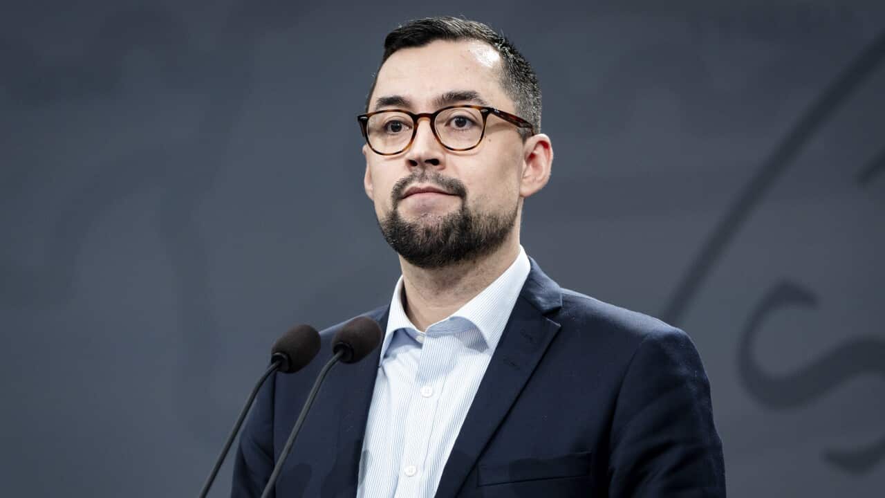 A man with a beard and glasses and wearing a suit and tie stands at a microphone