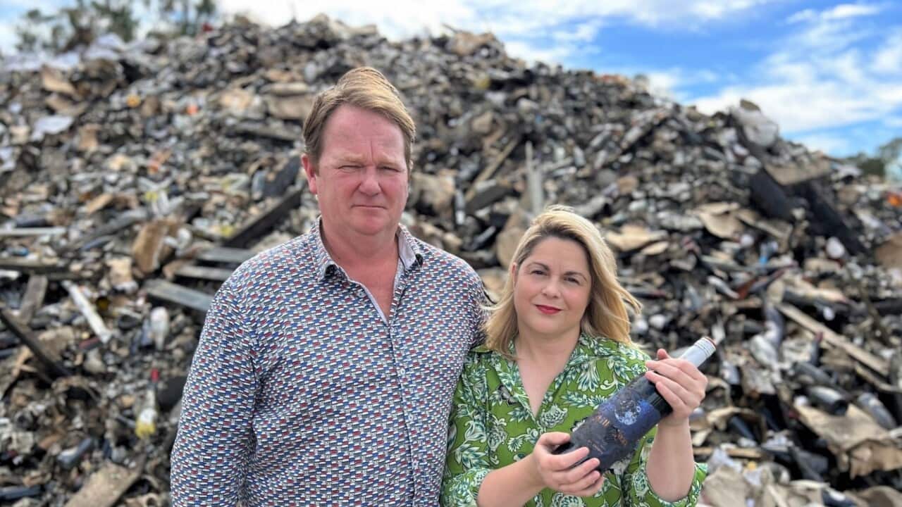 A man and a woman stand frowning in front of a pile of debris. The woman is holding a charred bottle of wine.