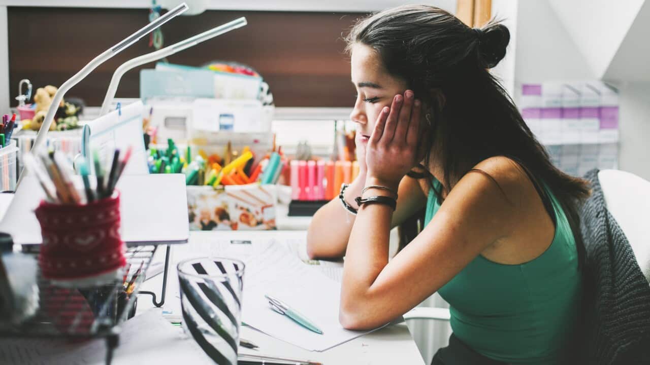 A female teenager sits at a desk with her hands on her face, looking at her notes. She is surrounded by stationery items.