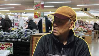 An elderly Japanese man wearing a black zip-up jumper and a baseball cap is shown in the foreground, overlaid on an image of a supermarket where shoppers are walking among the aisles with fresh vegetables.