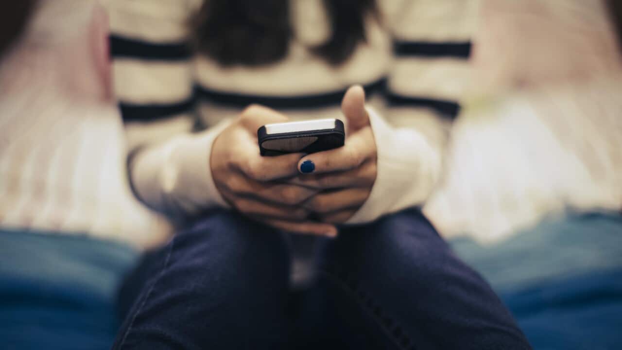 A close up of a teenager sitting on a bed using a smart phone