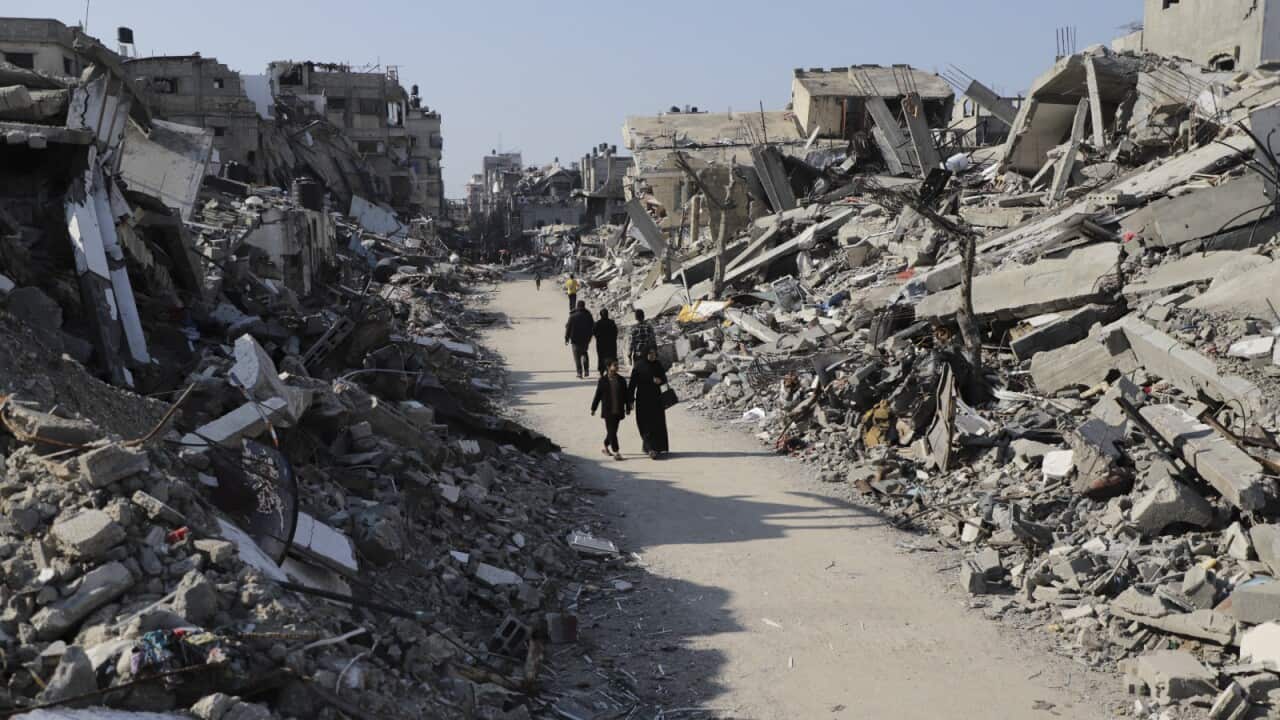 People walk through a cleared dirt road with collapsed buildings and rubbles surrounding them on either side.