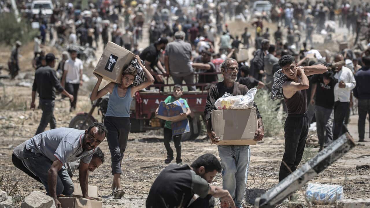 People are carrying cardboard boxes filled with humanitarian aid in a dry, grassy area.