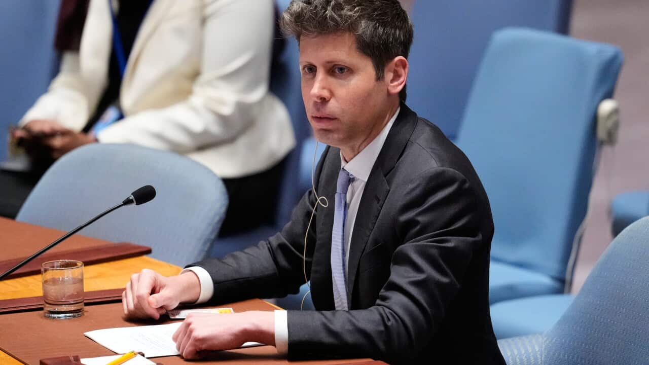 A man in a dark suit and pale blue tie sits at a wooden desk with a microphone and earpiece, looking off to the side with a serious expression.