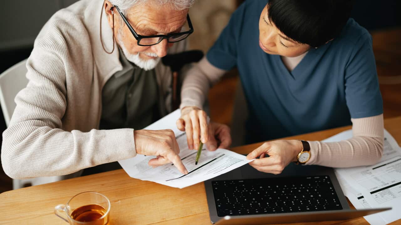 An elderly man wearing glasses looks at a document with a nurse wearing blue garb helping him read the paper. A cup of tea and a computer are also seen on the table.