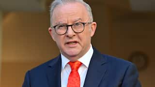 The image depicts Anthony Albanese at a press conference. The image is a medium close up. Albanese, a man in his 60s with greying hair, wears a navy blue business suit with a red patterned tie and round black glasses. He is speaking with some energy.
