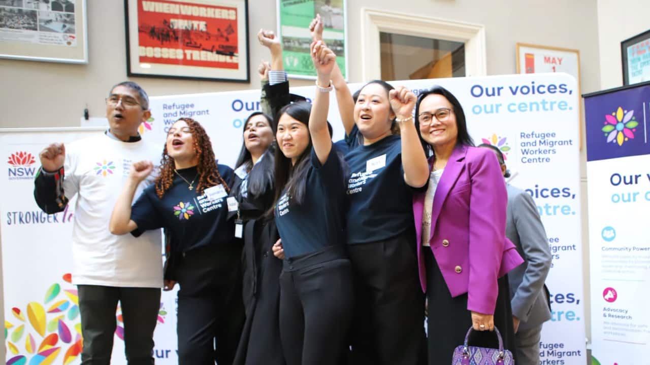 A diverse group of advocates stands together on a small stage, raising their fists and smiling during the launch of the Refugee and Migrant Workers Centre.