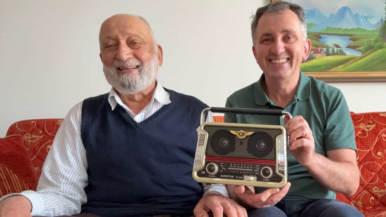 An elderly man on the left sits on a red couch with another man sitting and holding a radio on the right