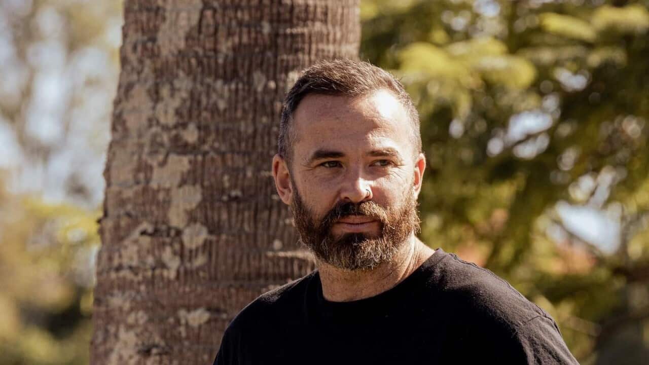 A man wearing a black shirt stares off into the distance. Trees are behind him.
