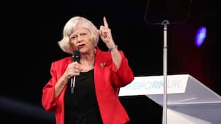 An older woman in a red coat and black blouse speaks into a microphone with her left hand raised.