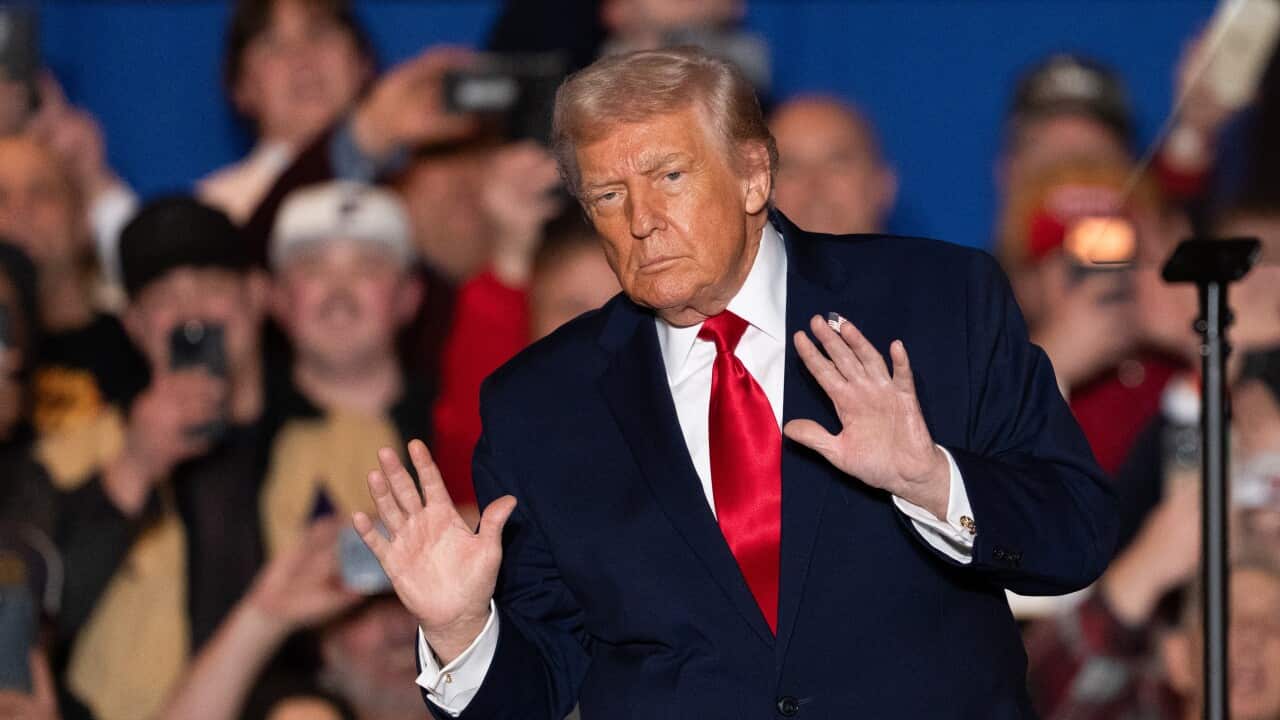 Donald Trump, with both hands raised, palms towards the crowd, with a serious look on his face, during a rally.