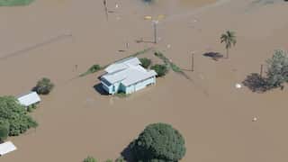 An aerial photo of a flooded home in Bundaberg, Queensland. The image features a solitary blue house with silver colourbond roof in the centre, its yard and the surrounding streets awash in brown floodwater.