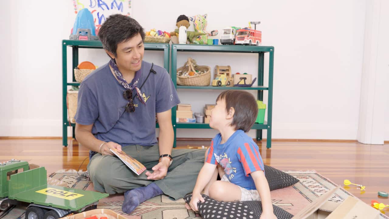 A man is sitting in a living room surrounded by toys playing with his son on the floor