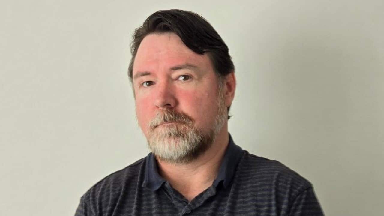 A man with a grey beard is posing in front of a white wall with a serious look on his face and his hands placed over one another