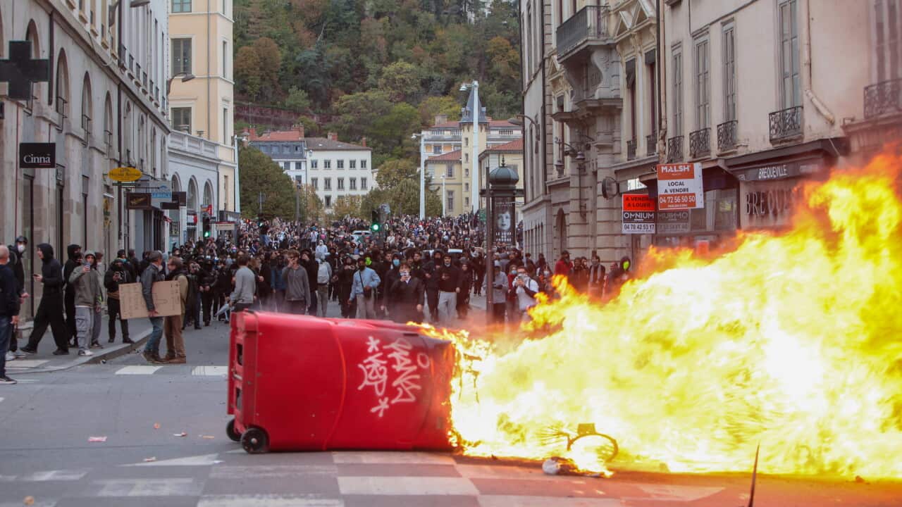 A large street protest fills a city road lined with multi-storey historic buildings. In the foreground, an overturned red rubbish bin lies on its side near a road intersection while a large fire burns across part of the street, producing bright yellow and orange flames. A dense crowd of hundreds of people stretches into the distance behind the fire, occupying the width of the roadway. Shopfronts, streetlights and apartment balconies line both sides of the street, and a tree-covered hillside rises in the background behind the urban streetscape. Visible signs on buildings include “MALSH”, “BREQUILLE” and a real estate advertisement.