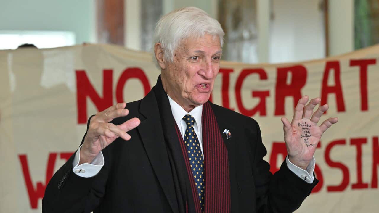 Bob Katter, an elderly man with white hair and wearing a black coat, gestures with his hands—one of which has writing on the palm—while standing in front of a banner reading "no migration without assimilation".